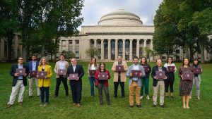 A group of people holding certificates stand on Killian Court in front of the MIT dome.